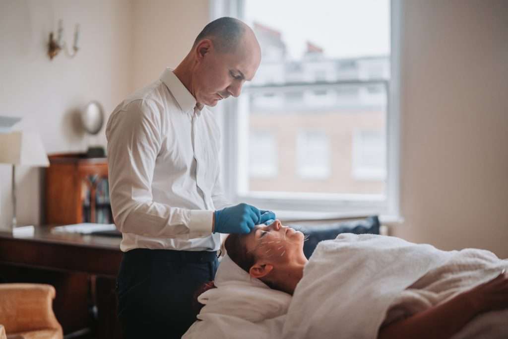 Dr John Tsagaris performing facial acupuncture treatment on a patient in a London consultation room overlooking Harley Street